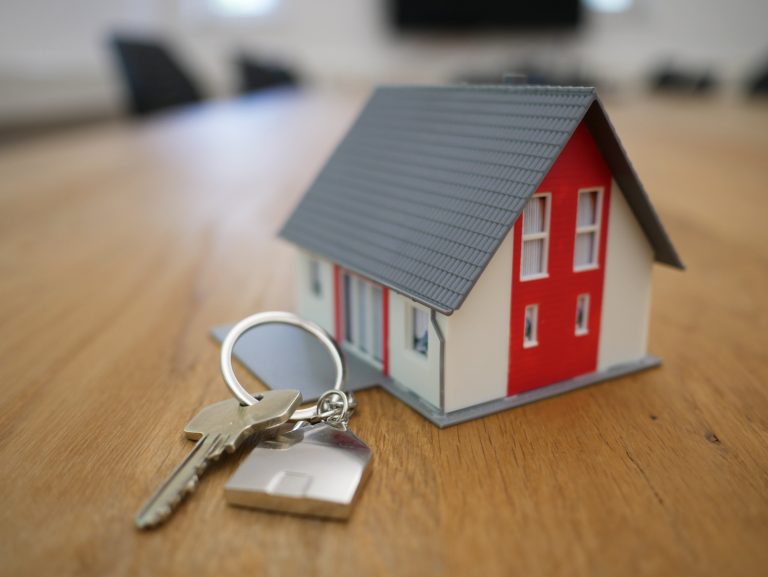 The steps to buying a house in Ireland. The image depicts a small model of a red and white house next to a key on a silver keychain.